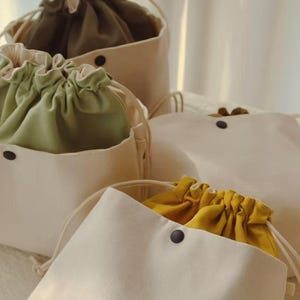 May include: A collection of off-white canvas drawstring pouches featuring interior linings in olive green, sage green, and mustard yellow. Each pouch has a black snap button closure on the front and a small white tag labeled FINESTRA on the side.