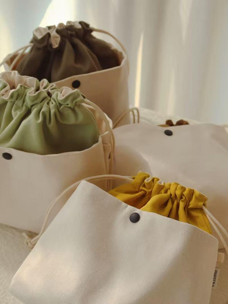 May include: A collection of off-white canvas drawstring pouches featuring interior linings in olive green, sage green, and mustard yellow. Each pouch has a black snap button closure on the front and a small white tag labeled FINESTRA on the side.
