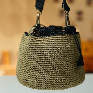 May include: A small, beige woven bucket bag with a black drawstring closure and strap. The bag has a rounded shape and is suspended by a black strap with silver-colored metal clasps. The bag's texture is visible, showcasing the woven pattern.