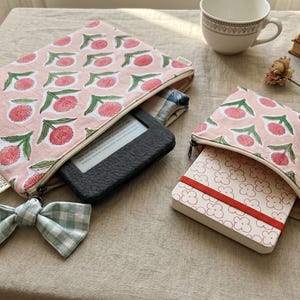 Könnte beinhalten: Zwei Stofftaschen mit Reißverschluss in Hellrosa mit einem sich wiederholenden Muster aus rosa Blumen und grünen Stielen. Die größere Tasche enthält einen schwarzen E-Reader und hat eine hellgrün-weiß karierte Schleife am Reißverschluss. Die kleinere Tasche enthält ein weißes Notizbuch mit rotem Gummiband.