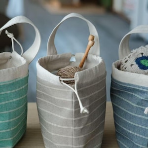 Könnte beinhalten: Drei gestreifte Stoffaufbewahrungstaschen mit Kordelzug. Die Taschen sind in Türkis, Grau und Blau. Die türkisfarbene Tasche ist leer. Die graue Tasche enthält Garn und eine Stricknadel aus Holz. Die blaue Tasche enthält eine gehäkelte Deckchen.