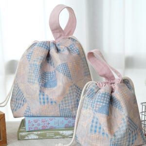 May include: Two drawstring pouches featuring a patchwork pattern of light blue and peach-colored squares with various geometric prints. Each bag has a solid pink fabric handle at the top and white cord drawstrings to cinch the opening closed.