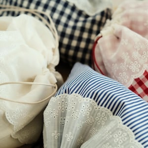 May include: Close-up of fabric bundles in various patterns and colours. Includes white, blue and white striped, red and white checked, and navy blue gingham fabrics. Some bundles have decorative lace trim and drawstring closures.
