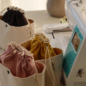 May include: Three canvas drawstring bags in black, mustard yellow, and dusty pink, each featuring a cream-colored exterior, a black snap button on the front, and a matching cream-colored drawstring closure, positioned next to a white sewing machine.