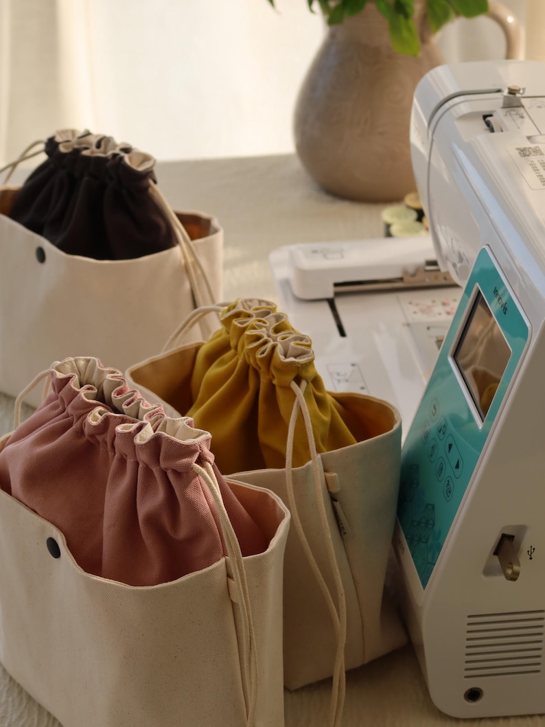 May include: Three canvas drawstring bags in black, mustard yellow, and dusty pink, each featuring a cream-colored exterior, a black snap button on the front, and a matching cream-colored drawstring closure, positioned next to a white sewing machine.