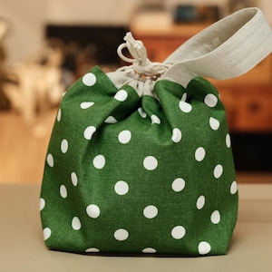 May include: A green drawstring bag with white polka dots. The bag has a natural-colored linen handle and drawstring closure. The bag is made of fabric and is sitting on a neutral surface.