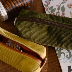 May include: An open yellow corduroy pencil case with a brown zipper and a comb inside. A closed green corduroy pencil case with a brown zipper is next to it. A jar of cookies and a floral print card are also in the image.