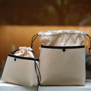 Pode incluir: Duas bolsas de tecido bege com acabamento preto e cordões. A bolsa maior tem um detalhe de botão preto. As bolsas são de tamanhos diferentes e têm uma aparência macia e texturizada. O fundo está desfocado, sugerindo um ambiente interno.