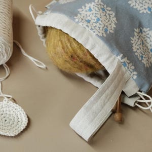 May include: A craft scene with a white yarn spool, a crocheted circle, and a blue floral drawstring bag containing a ball of yarn. Knitting needles are visible inside the bag. The items are arranged on a neutral surface.