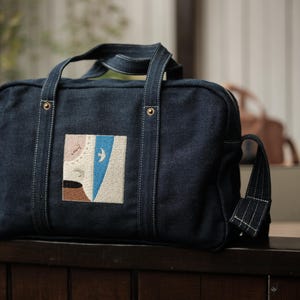 May include: A dark blue denim rectangular bag featuring two top handles and a side strap. The front of the bag displays a square patch with an embroidered abstract design in shades of beige, brown, and blue, including a small white bird icon.