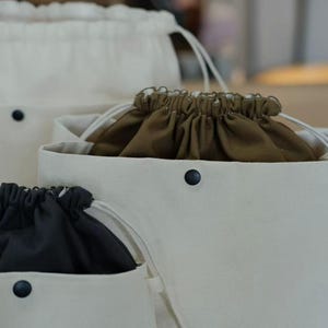 Puede incluir: Tres bolsas de lona con cordón en diferentes tamaños. Las bolsas son de color blanquecino con detalles y cordones negros. El interior de cada bolsa es de un color diferente, incluyendo negro y verde oliva.