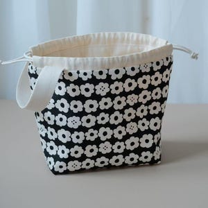 May include: A small tote bag with a black floral pattern on a white background. It features a white drawstring closure and handle. The interior is a plain off-white colour.