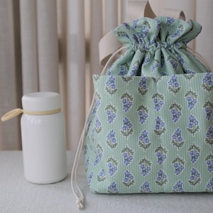 May include: A white insulated water bottle with a tan loop handle sits beside a drawstring bag. The bag is light green with a floral pattern in blue and green. The bag has a beige drawstring and a beige ribbon handle.