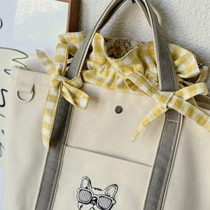 Puede incluir: Un bolso tote de lona color crema con correas verticales grises, un bolsillo frontal con un bulldog francés bordado con gafas de sol y un cierre de cordón de cuadros vichy amarillos y blancos con dos lazos decorativos.