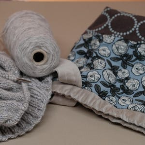 May include: A still life image featuring knitting supplies. A large spool of gray yarn sits next to a partially knitted gray scarf. A fabric piece with a floral pattern in blue and white is also present, suggesting a crafting theme.