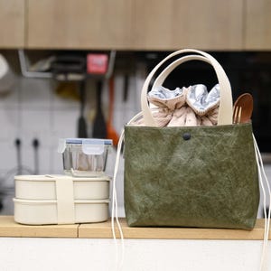 May include: A green tote bag with cream-colored handles and drawstring closure, holding a patterned lunch bag and wooden spoon. Beside it are two stacked beige lunch boxes with a clear glass container on top.