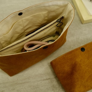 May include: A brown suede tote bag with a matching clutch. The tote bag has a zipper closure and a light-colored interior with pockets. The clutch has a single black snap closure. The bags are on a light gray surface.