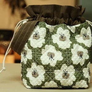 Puede incluir: Bolso con cordón acolchado con un estampado floral. El bolso presenta flores blancas sobre un fondo verde, un cierre de cordón marrón y una correa para el hombro a juego. El bolso mide aproximadamente 20 cm de alto.