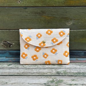 Könnte beinhalten: Eine beige Clutch mit Blumenmuster und Klappenverschluss. Die Clutch hat ein Muster aus orangefarbenen Blumen mit blauen Akzenten. Sie wird mit einem goldfarbenen Druckknopf verschlossen. Der Hintergrund ist eine verwitterte Holzoberfläche.