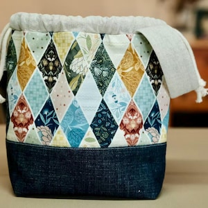 May include: A fabric storage bag with a denim base and a patterned upper section. The upper section features a diamond pattern in various colors, including blue, yellow, and green. A drawstring closure and a white fabric trim complete the design.
