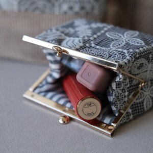 Peut inclure: Un petit porte-monnaie rectangulaire dot&eacute; d'un fermoir m&eacute;tallique dor&eacute;. L'ext&eacute;rieur est en tissu noir et blanc recouvert d'une dentelle florale blanche. &Agrave; l'int&eacute;rieur, un rouge &agrave; l&egrave;vres rouge et un produit cosm&eacute;tique carr&eacute; mauve sont visibles.