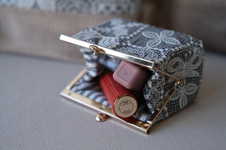 Peut inclure: Un petit porte-monnaie rectangulaire dot&eacute; d'un fermoir m&eacute;tallique dor&eacute;. L'ext&eacute;rieur est en tissu noir et blanc recouvert d'une dentelle florale blanche. &Agrave; l'int&eacute;rieur, un rouge &agrave; l&egrave;vres rouge et un produit cosm&eacute;tique carr&eacute; mauve sont visibles.
