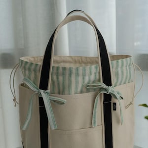 Op de afbeelding: Een beige tote bag met zwarte bandjes en een gestreepte binnenkant. De tas heeft twee buitenvakken en is versierd met lichtgroene strikken. De handvatten zijn beige met zwarte accenten.