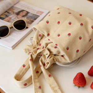 Pode incluir: Bolsa bege com cordão de veludo cotelê com um padrão repetido de morangos vermelhos. A bolsa tem uma parte superior com babados e alças longas. Óculos de sol e uma revista são visíveis no fundo. Também há morangos.