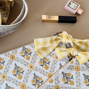 May include: A rectangular pouch featuring a white fabric with a yellow and dark green floral pattern, accented by two yellow and white gingham bows attached to the zipper pull. A matching yellow gingham bag handle is visible in the upper left corner.