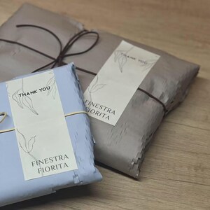 May include: Two rectangular packages wrapped in light blue and taupe paper, secured with thin dark string. Each package features a cream-colored label with a line-art leaf illustration, the text THANK YOU, and the brand name FINESTRA FIORITA.