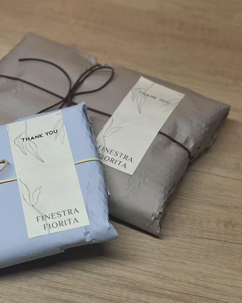 May include: Two rectangular packages wrapped in light blue and taupe paper, secured with thin dark string. Each package features a cream-colored label with a line-art leaf illustration, the text THANK YOU, and the brand name FINESTRA FIORITA.