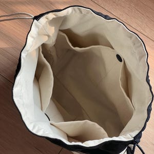 May include: A black and off-white drawstring bag with a wide opening, featuring a cream-colored interior lining, multiple internal slip pockets, and a black drawstring cord closure that cinches the top of the bag securely.