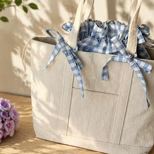 May include: A beige canvas tote bag featuring a blue and white gingham patterned drawstring closure at the top. The bag includes two sturdy canvas handles and a vertical stitched pocket detail on the front, with blue gingham fabric ties.