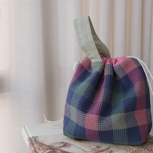 以下が含まれることがあります： 青、ピンク、緑の色合いのカラフルなチェック柄の小さな巾着袋。 バッグにはライトグレーのハンドルとピンクの裏地が付いています。 白いドローストリングコードが見えます。