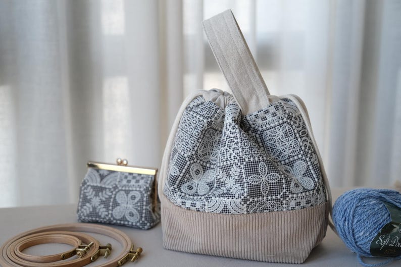 Peut inclure: Un sac bourse &agrave; cordon avec une base en velours c&ocirc;tel&eacute; beige et une partie sup&eacute;rieure en tissu noir et blanc &agrave; motif dentelle. Le sac poss&egrave;de une anse beige unique et un petit porte-monnaie assorti avec un cadre et fermoir en m&eacute;tal dor&eacute;.