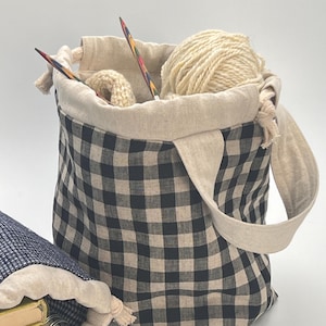 May include: A black and cream checked tote bag with a natural linen lining and handle. The bag contains yarn, knitting needles, and a small knitted item. A second bag with books is visible in the background.