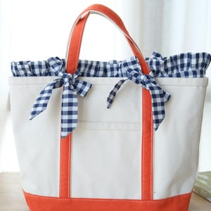 May include: A cream-colored canvas tote bag featuring bright orange trim on the base and handles. The top opening is lined with a navy blue and white gingham fabric that includes two decorative bows tied at the front of the bag.
