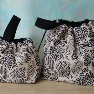 May include: Two drawstring pouches in a beige fabric with a black animal print pattern. Each bag features a black fabric handle and black drawstring cords. The pouches are of different sizes, with the smaller one on the left and the larger one on the right.