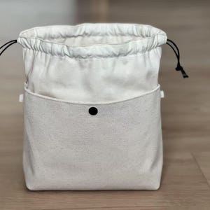 Puede incluir: Una bolsa con cordón de color crema con un bolsillo delantero y un cierre de botón a presión negro. La bolsa tiene cordones negros y está hecha de una tela texturizada. La bolsa está diseñada para mantenerse de pie y probablemente se utiliza para el almacenamiento.
