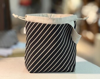 Pode incluir: Uma bolsa com cordão de riscas pretas e brancas. A bolsa apresenta um tecido preto com riscas diagonais brancas. A borda superior é de cor bege claro, e o cordão é bege a condizer. A bolsa está sobre uma superfície branca.