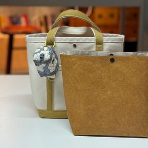 May include: Two tote bags on a white surface. One is off-white with olive green accents and a small patterned charm. The other is a brown paper bag style tote. Both bags are empty.