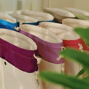 May include: A collection of drawstring pouches featuring a two-tone design with a cream-colored base and a contrasting upper section in vibrant colors including purple, lavender, red, and blue, each with a white drawstring cord and a snap button.
