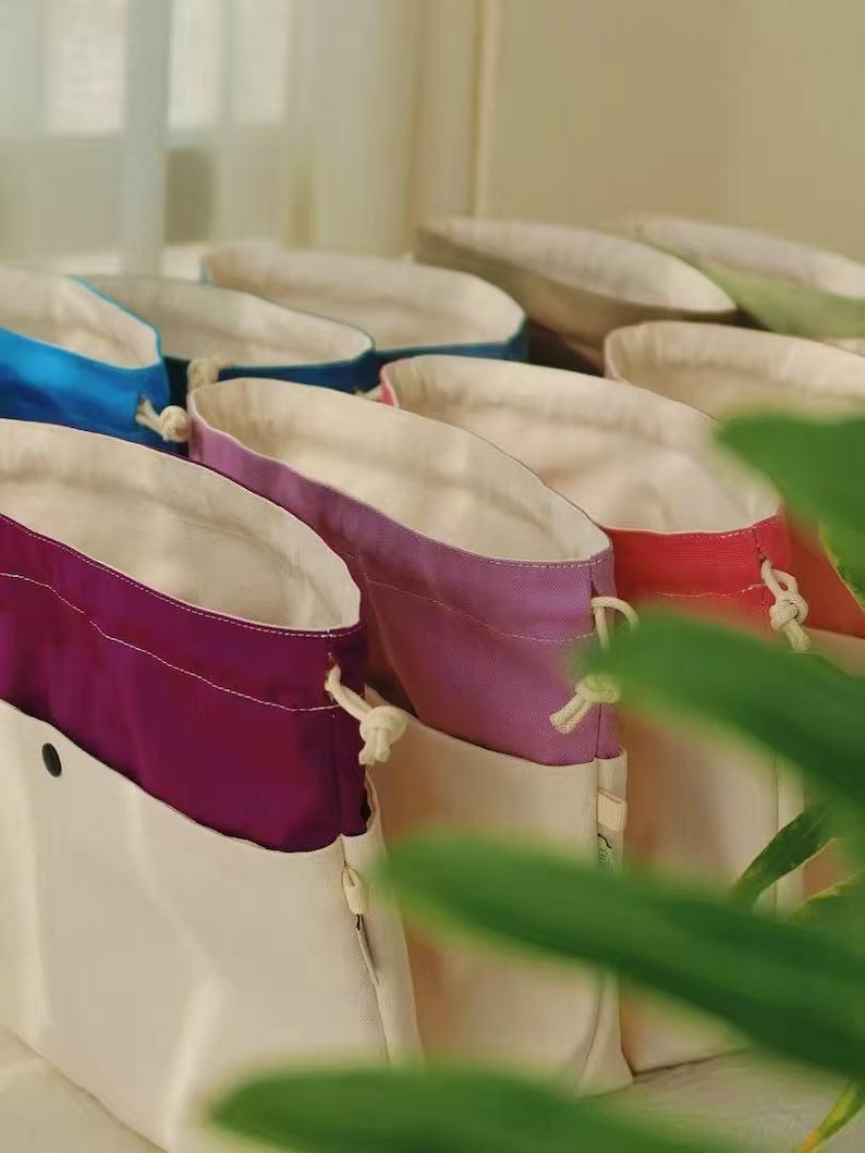 May include: A collection of drawstring pouches featuring a two-tone design with a cream-colored base and a contrasting upper section in vibrant colors including purple, lavender, red, and blue, each with a white drawstring cord and a snap button.