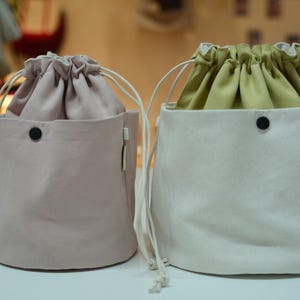 May include: Two drawstring bags. One is a light pink color, and the other is off-white. Both have a cylindrical shape with a drawstring closure and a black button detail. The interior of the off-white bag is olive green.