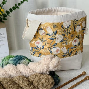 May include: A yellow and white floral print yarn storage bag with a cream-coloured canvas base and handle. The bag contains various colours of yarn and two wooden knitting needles. The bag is designed for storing yarn and knitting supplies.