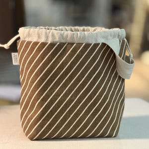 May include: A brown and white striped drawstring bag with a light-colored linen top and handle. The bag has a square base and is made of fabric with diagonal stripes. A small tag with text is visible on the side.
