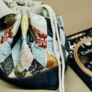 May include: A patchwork drawstring bag with a denim base and a gray fabric top. The bag features a diamond pattern in various colors and floral designs. A book and embroidery hoop are in the background.