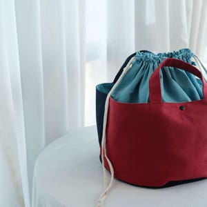 Peut inclure: Deux sacs fourre-tout en toile. Le sac avant est rouge avec un intérieur bleu et des poignées. Le deuxième sac est bleu marine avec un intérieur bleu. Les deux sacs ont des fermetures à cordon et sont sur une surface blanche.