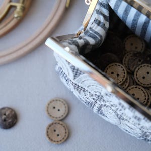 Peut inclure: Une petite pochette en tissu avec un fermoir m&eacute;tallique &agrave; cliquet, dot&eacute;e d'un ext&eacute;rieur &agrave; motifs noirs et blancs et d'une doublure int&eacute;rieure ray&eacute;e noire et blanche. La pochette est remplie de nombreux boutons en bois clair &agrave; deux trous.