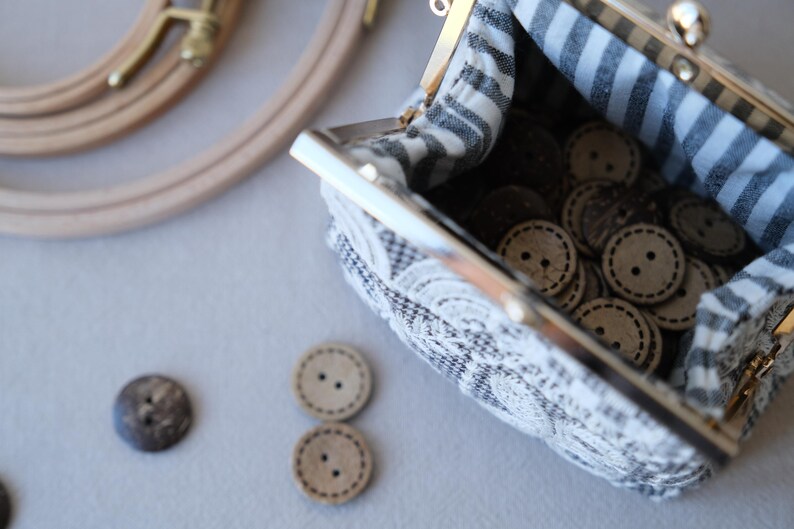 Peut inclure: Une petite pochette en tissu avec un fermoir m&eacute;tallique &agrave; cliquet, dot&eacute;e d'un ext&eacute;rieur &agrave; motifs noirs et blancs et d'une doublure int&eacute;rieure ray&eacute;e noire et blanche. La pochette est remplie de nombreux boutons en bois clair &agrave; deux trous.